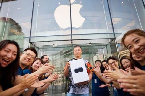 من iPhoneIslam.com، رجل يقف حاملاً صندوقًا يحتوي على Apple Vision Pro خارج متجر Apple Store في يوليو، محاطًا بمجموعة من الأشخاص يصفقون ويبتسمون.