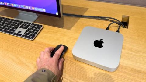 من iPhoneIslam.com، لقطة مقربة ليد شخص يستخدم ماوس لاسلكي بجوار جهاز Mac mini وشاشة خارجية على مكتب خشبي.