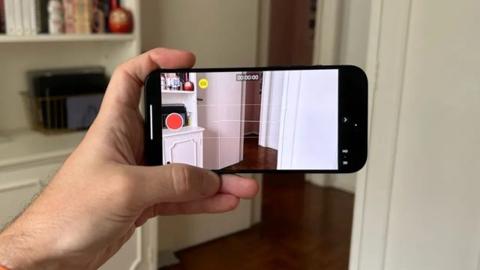 من iPhoneIslam.com، شخص يحمل هاتفًا ذكيًا لالتقاط فيديو لغرفة باستخدام ميزة الالتقاط المكاني.