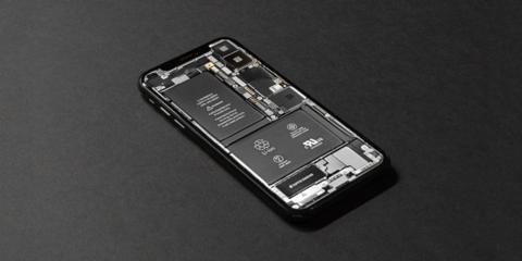 من iPhoneIslam.com، صورة مقربة لبطارية iPhone على سطح أسود، مع نصائح لإطالة عمر البطارية.