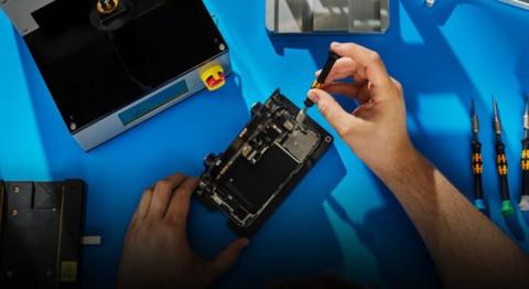 من iPhoneIslam.com، يقوم شخص بتفكيك أو إصلاح جهاز الآي فون الموجود على مكتب مع وجود أدوات ومعدات متناثرة حوله.