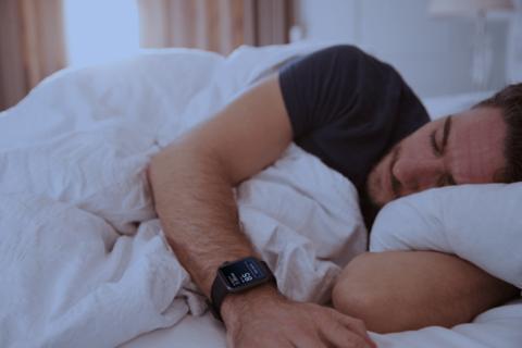 من iPhoneIslam.com، شخص يرتدي ساعة Garmin الذكية يرقد في السرير تحت لحاف أبيض، ويبدو أنه نائم.