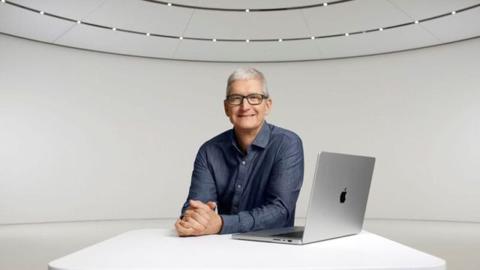 من iPhoneIslam.com، يجلس تيم كوك في شركة Apple على طاولة مع جهاز كمبيوتر محمول.