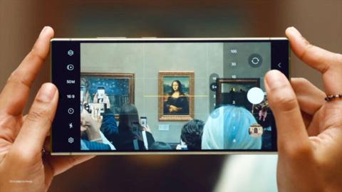 من iPhoneIslam.com، شخص يستخدم هاتفًا ذكيًا من سلسلة Galaxy S24 لالتقاط صورة للوحة فنية.